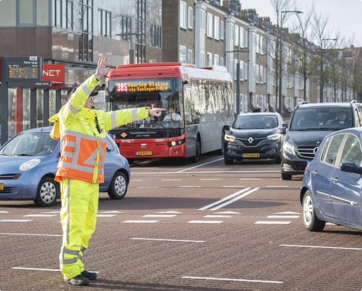 Afbeelding van een verkeersregelaar in fel reflecterende werkkleding aan het werk op een druk kruispunt met auto's en een stadsbus